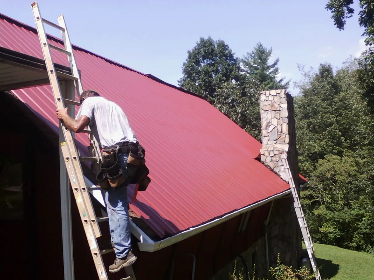 Roofer on a ladder beside a red metal roof and stone chimney — Soffit & Fascia Repair by Roofing Brothers in Castroville